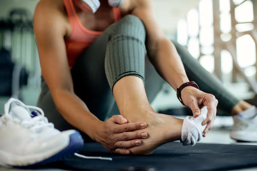 Individual sitting on a black exercise mat, showing their foot and ankle near gym shoes in a physical therapy context.