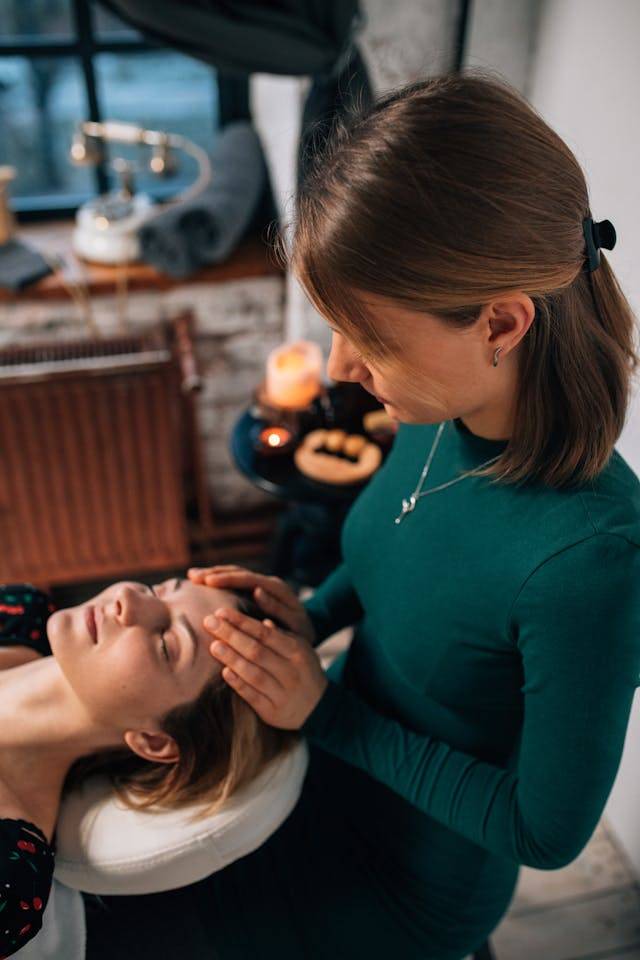 Two women providing a facial treatment with tools and candles in a cozy setting.
